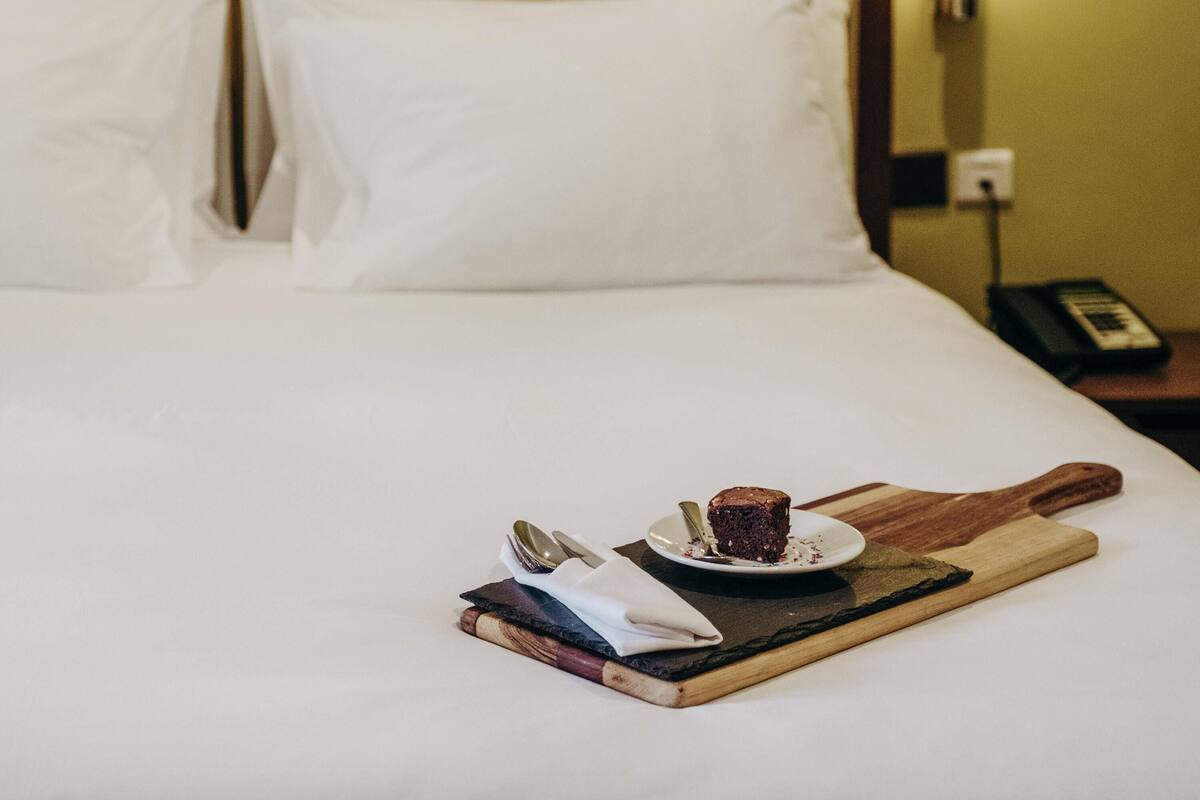 Cama bem arrumada com lençóis brancos, mesa de cabeceira com telefone e bandeja de madeira com fatia de bolo e talheres.