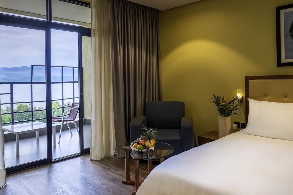 Quarto de hotel com uma cama grande, uma cadeira, uma mesinha com frutas, um vaso de planta e uma varanda com vista.