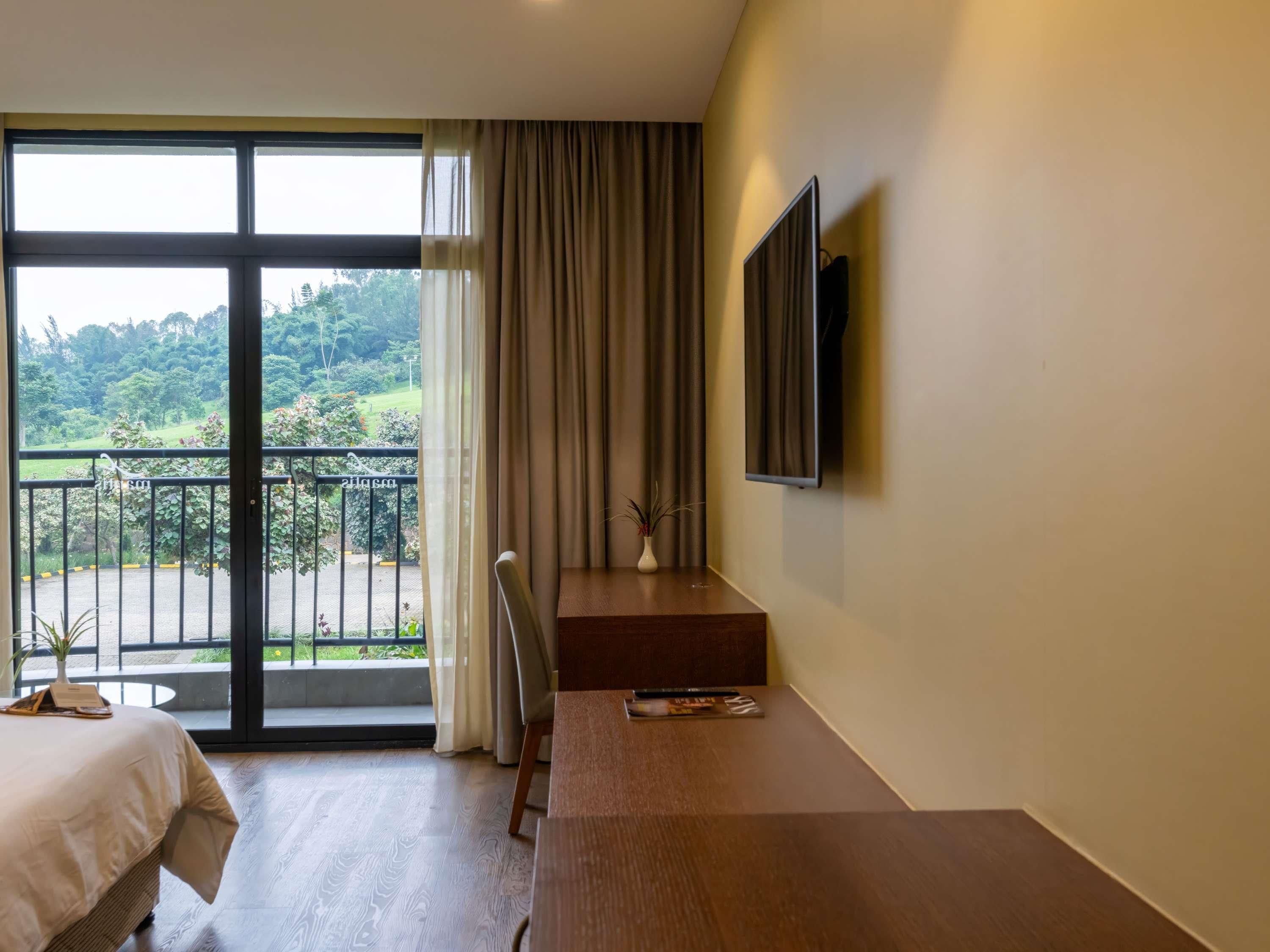A hotel room with a bed, a desk, a chair, a TV, and a balcony with a view of greenery.