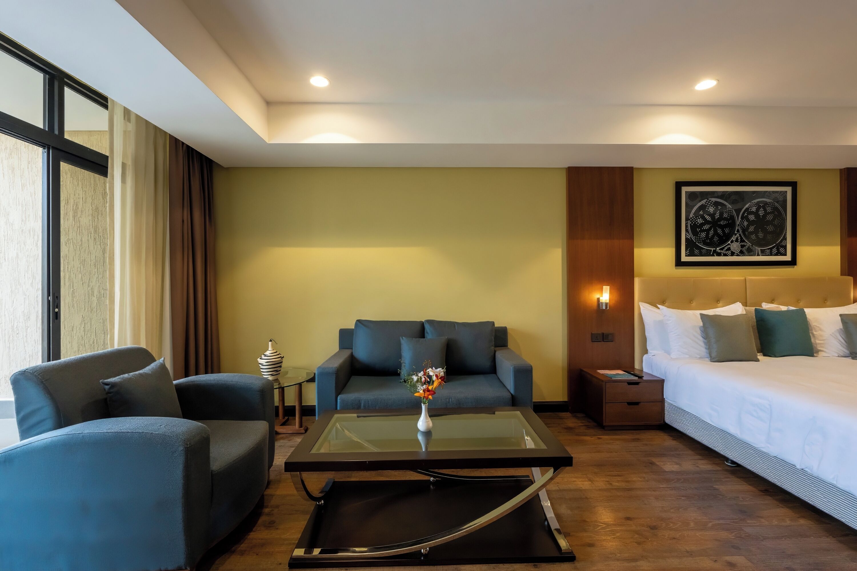 A modern hotel room with a sofa, a glass coffee table, a bed, and a nightstand.