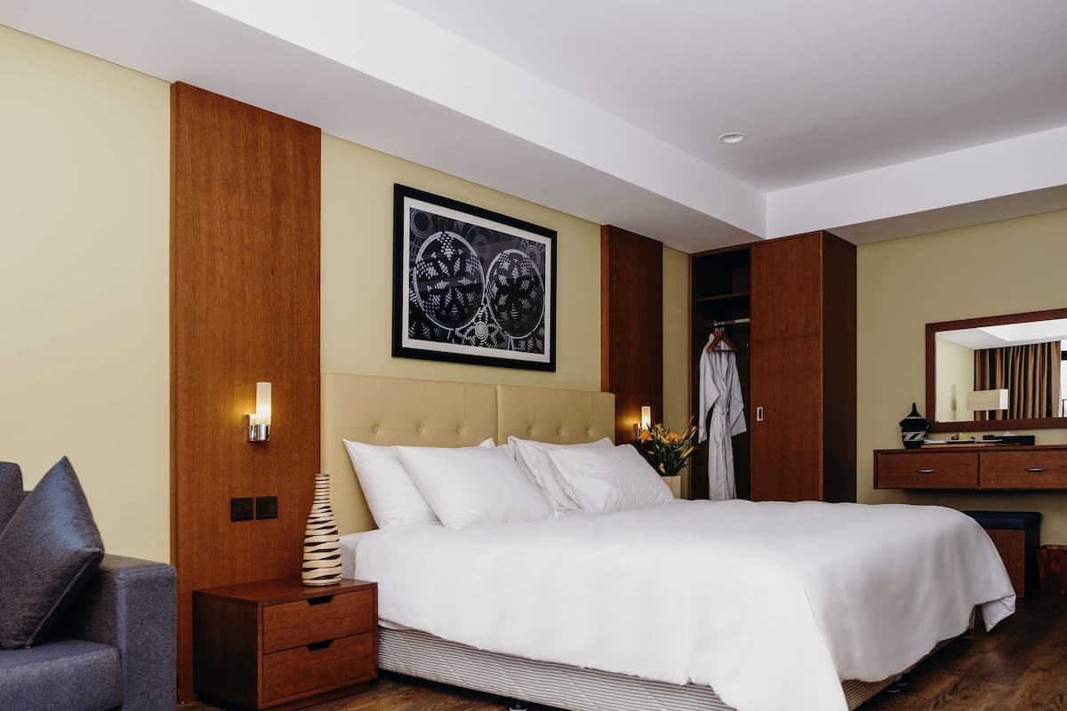 Quarto de hotel com uma cama grande, mesa de cabeceira, luminária, cadeira, guarda-roupa e espelho.
