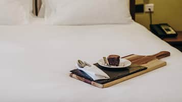 Una cama bien tendida con ropa blanca, una mesita de noche con un teléfono y una bandeja de madera con una porción de pastel y cubiertos.