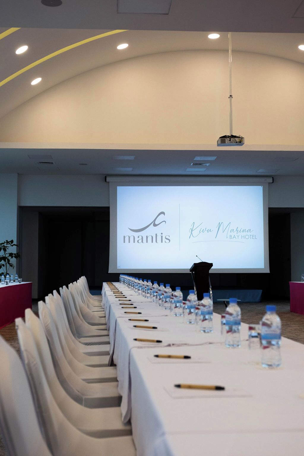 Un salón de conferencias con una larga mesa dispuesta para una reunión, un atril y una pantalla mostrando el logotipo y el nombre del Hotel Kiwa Marina.