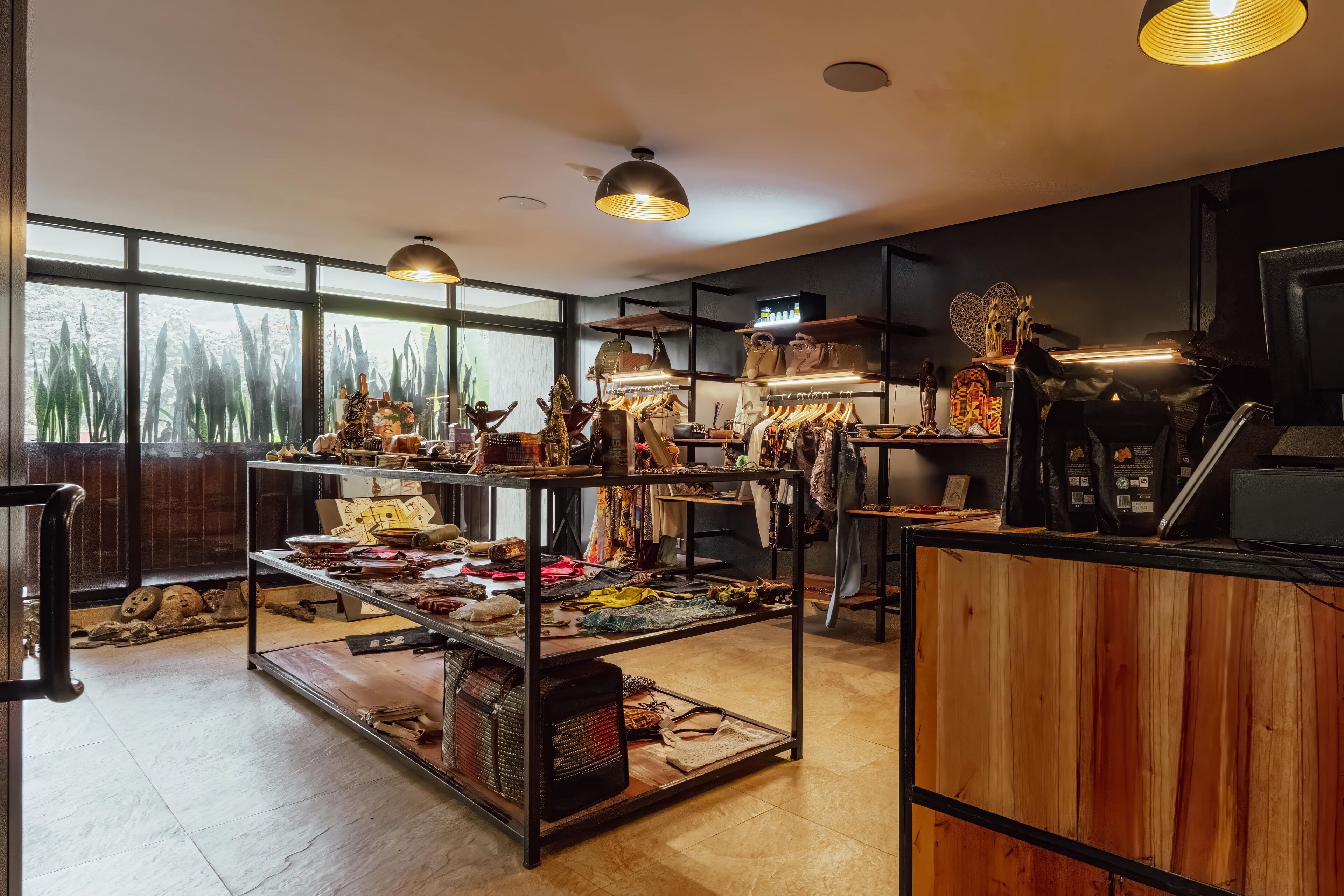 A boutique store with shelves of clothing and accessories, wooden flooring, and large windows with a view of trees.