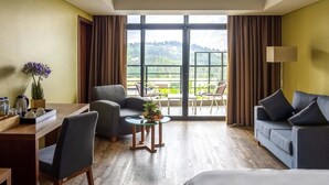 Una habitación de hotel con cama, sofá, mesa de comedor con sillas, una mesita con planta y vistas a un balcón con barandilla.