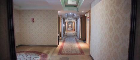 Ein Hotelgang mit gemustertem Teppich, einer Deckenleuchte und einem Feuermelder an der Wand.