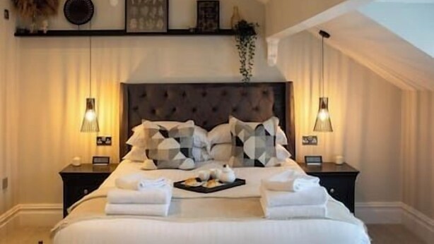 A neatly made bed with pillows and folded towels, a bedside table with a tray of food, and a wall-mounted shelf with decorative items. - King Suite close to Manchester City Centre, R3 (Stockport)