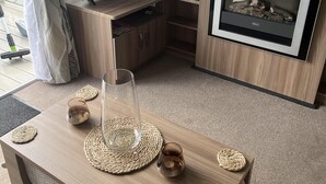 A modern living room with a wooden TV stand, a glass coffee table, and a fireplace. - Tranquil Waterside Lodge in Tunstall (Hull)