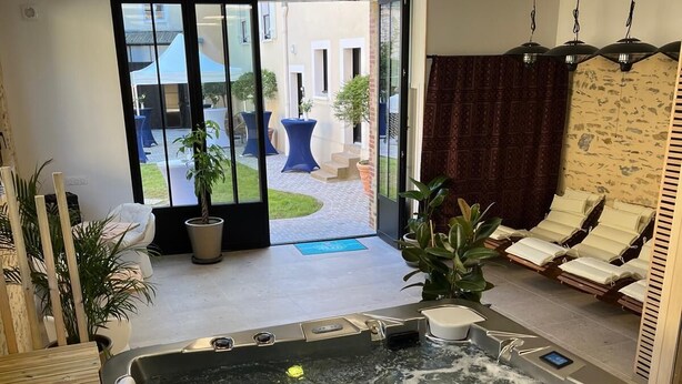 A modern indoor hot tub with a wooden deck, a glass door leading to an outdoor patio, and a seating area with lounge chairs. - Chambre Frida Kalho (Laval)