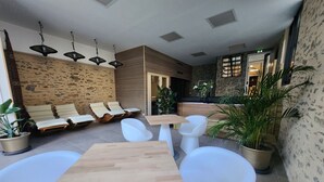 A modern outdoor seating area with wooden tables and white chairs, surrounded by stone walls and potted plants. - Chambre Frida Kalho (Laval)