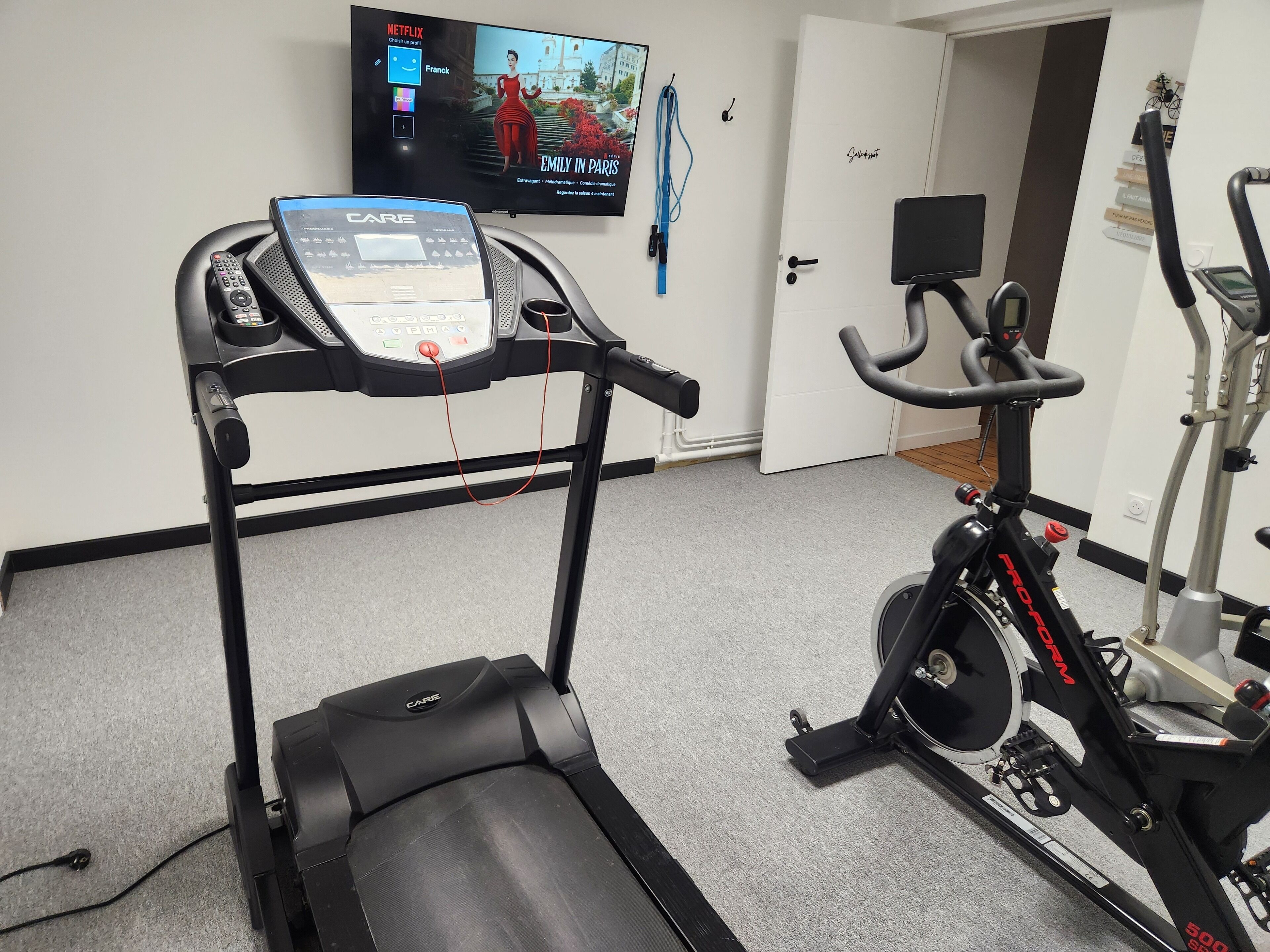 Une salle de sport à domicile équipée d’un tapis de course, d’un vélo d’appartement et d’un téléviseur diffusant un film.