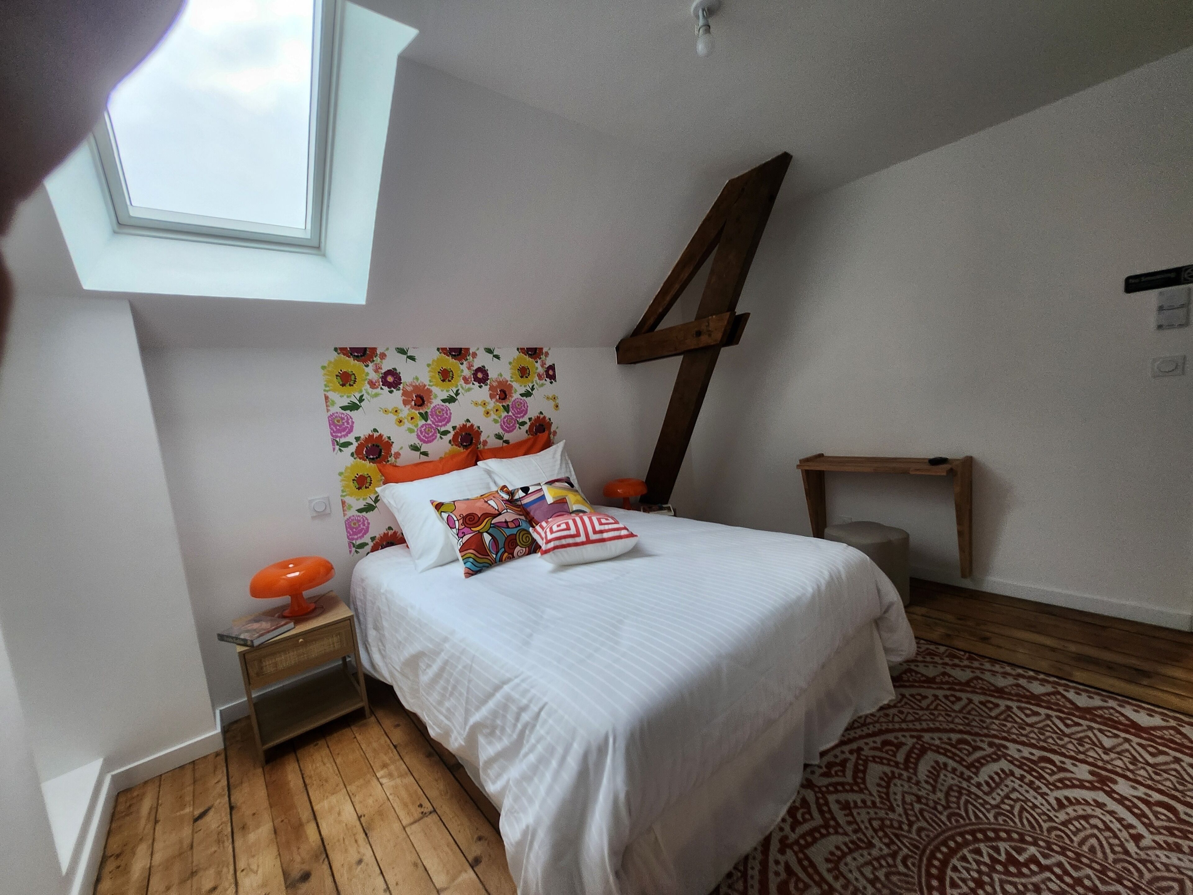 Une chambre avec un puits de lumière, un lit avec une literie blanche et des coussins colorés, un plancher en bois et un tapis à motifs.