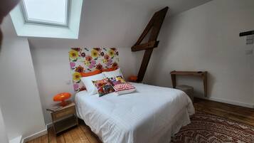 Une chambre avec un puits de lumière, un lit avec une literie blanche et des coussins colorés, un plancher en bois et un tapis à motifs.