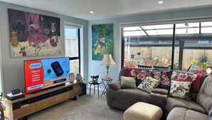 Ein Wohnzimmer mit einem großen Flachbildfernseher, einer Holzunterhaltungsecke, einem braunen Sofa und einem großen Fenster mit Blick auf einen Holzzaun und Pflanzen.