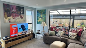 A living room with a large flat-screen TV, a wooden entertainment unit, a brown sofa, and a large window with a view of a wooden fence and plants. - A piece of Bali (Tauranga)