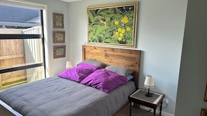 A bedroom with a bed, purple and gray bedding, a wooden headboard, a nightstand with a lamp, and a large framed painting above the bed. - A piece of Bali (Tauranga)