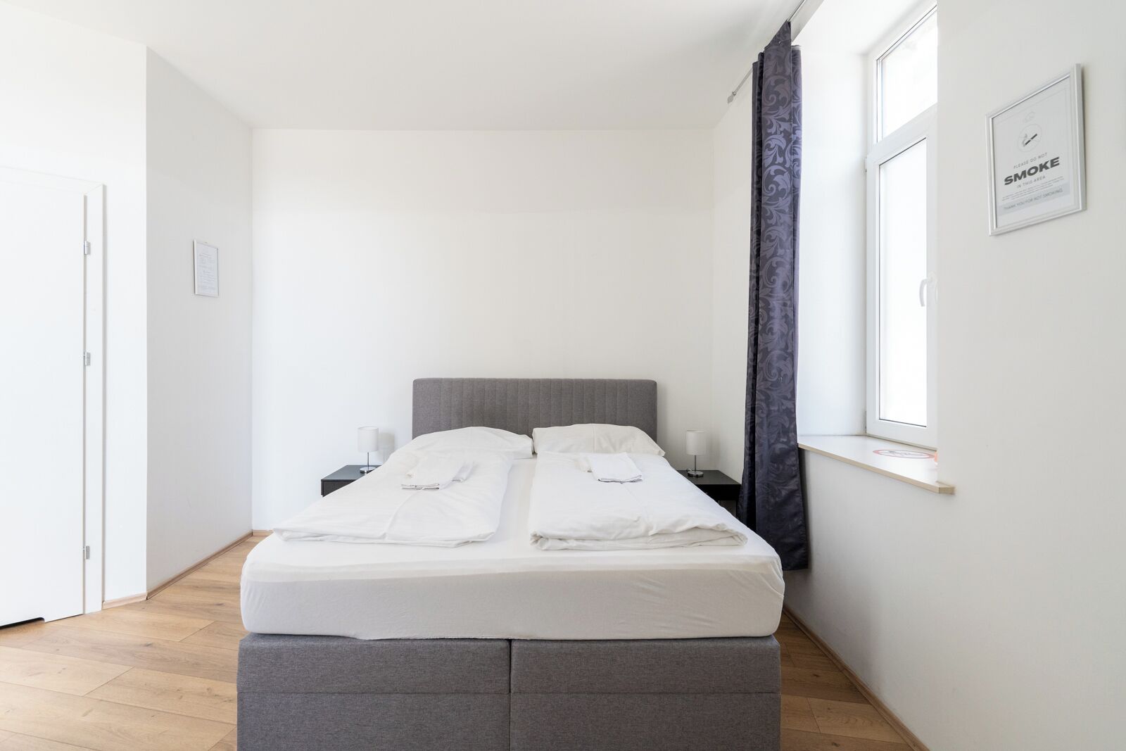A neatly made bed with white linens, a grey headboard, and a bedside table with a lamp.
