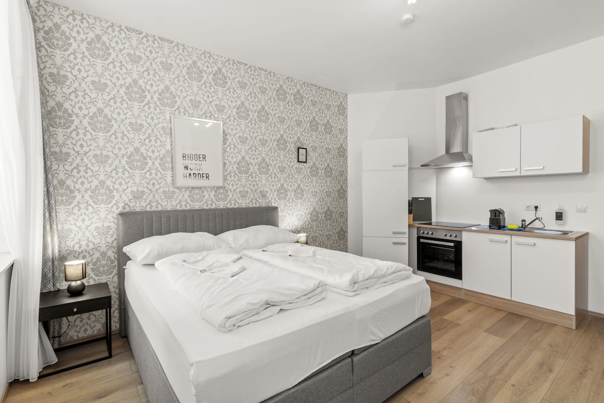 Un dormitorio moderno con una cama grande, una mesita de noche con lámpara y un pequeño tocador. Junto al dormitorio se encuentra una kitchenette con estufa, horno y armarios.