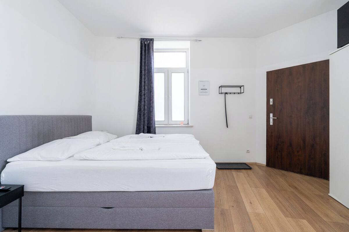 Un dormitorio limpio y moderno con una cama individual, cabecera gris, suelo de madera, ventana con cortinas, purificador de aire de pared y una puerta de madera oscura.