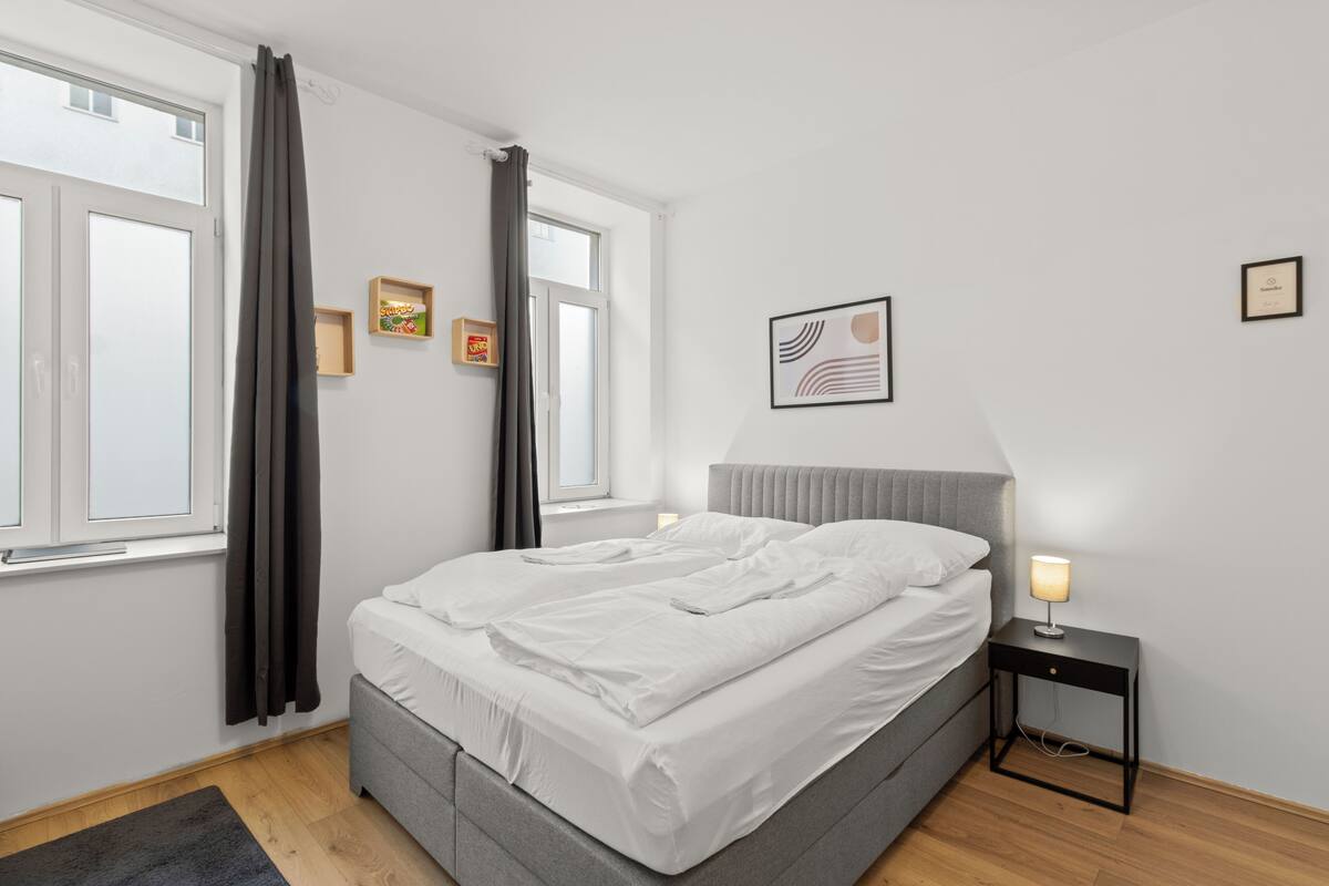 Un dormitorio moderno con una cama gris, ropa de cama blanca, una mesita de noche con lámpara y una ventana con cortinas.