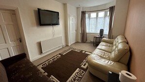 Una sala de estar con un sofá beige, un televisor de pantalla plana montado en la pared, un radiador, un tapete estampado, una ventana con cortinas y una pequeña mesa con un calentador.