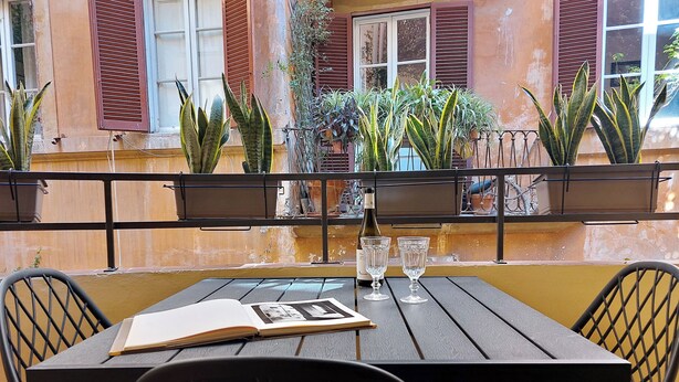 Una mesa con dos sillas, un libro y dos vasos en un balcón con plantas en macetas y barandilla.