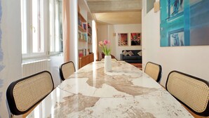 Un comedor con una mesa de mármol, sillas de mimbre, un jarrón con flores y estanterías al fondo.