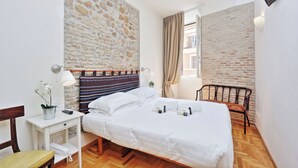 A hotel room with a stone wall, a bed with white linens, a bedside table with a lamp, a chair, and a TV mounted on the wall. - Spagna Charme - One Bedroom Apartment, Sleeps 4 (Rome)