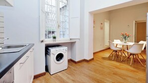 Une cuisine compacte dotée d’un lave-linge intégré, un coin repas avec une table et des chaises, et une fenêtre ornée de barreaux en fer décoratifs.