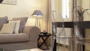 A modern living room with a sofa, a coffee table with books, a lamp, and clear plastic chairs.