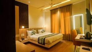A modern hotel room with a large bed, bedside table, and a desk with a chair. - Familia Hotel and Banquets (Lucknow)