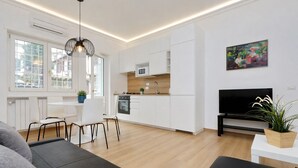 A modern kitchen with white cabinets, a dining area with a white table and chairs, a black sofa, and a TV on the wall. - Cozy Domar - Two Bedroom Apartment, Sleeps 6 (Rome)