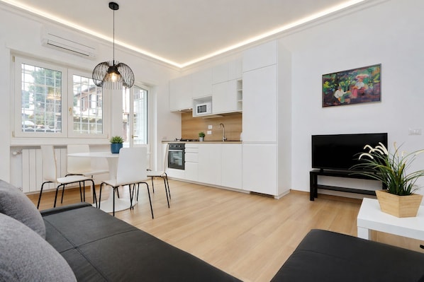 Una cocina moderna con armarios blancos, un comedor con mesa y sillas blancas, un sofá negro y un televisor en la pared.