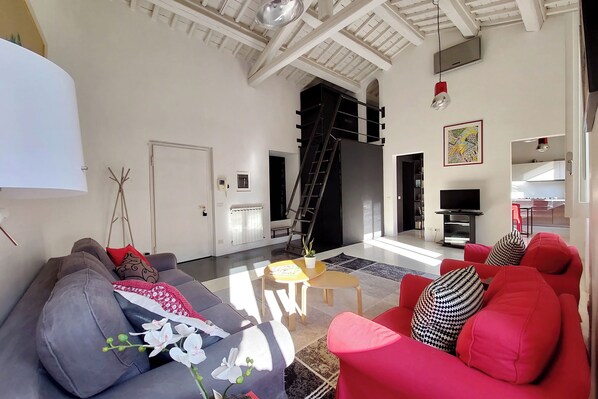 A spacious living room with a grey sofa, red armchair, and a small table with a plant. A staircase leads to an upper level, and a TV is mounted on the wall.
