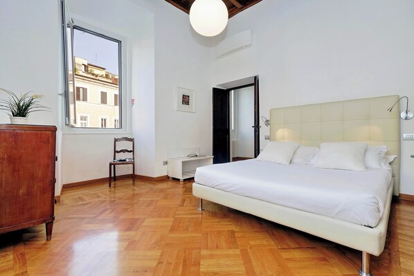 A bedroom with a large bed, wooden flooring, a window, a dresser, and a chair.