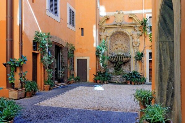 An outdoor courtyard with a cobblestone path, potted plants, and a fountain.
