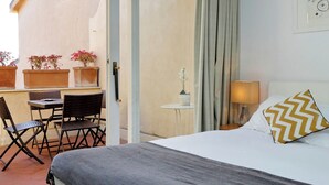 Habitación de hotel con una cama, una mesita con sillas, una planta en maceta, una lámpara y vistas al exterior.
