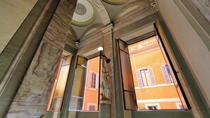 Habitación con techo alto, molduras ornamentales y vista a un edificio con contraventanas a través de una ventana.