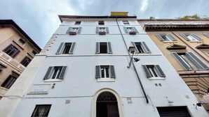 Ein mehrstöckiges Gebäude mit weißer Fassade, zahlreichen Fenstern mit Fensterläden und einem zentralen Eingang mit einer Tür.