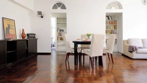Un comedor con mesa y sillas, una estantería y un sofá en una habitación con suelo de madera y paredes blancas.