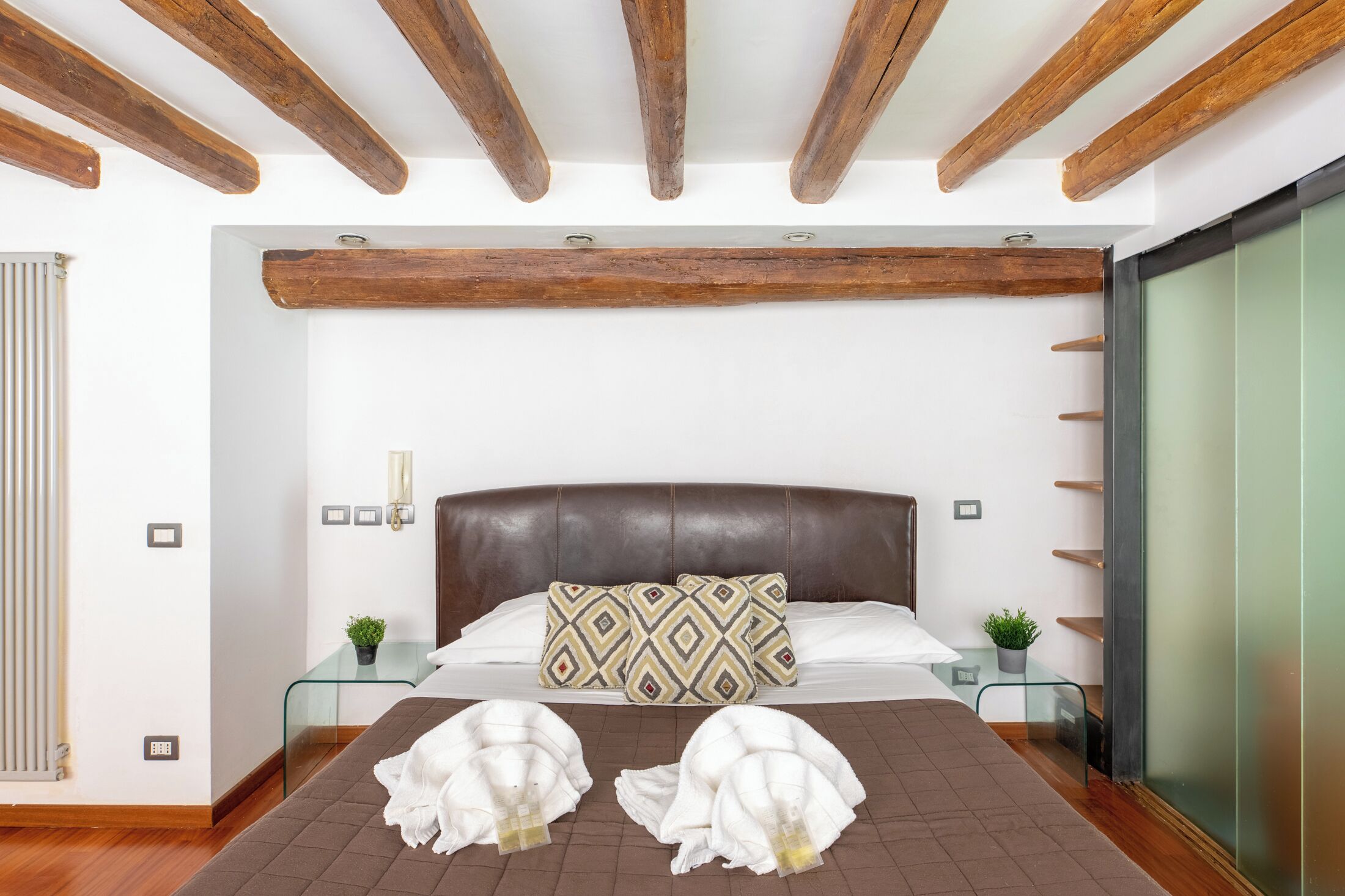 Une chambre avec un plafond en bois, un grand lit agrémenté d’oreillers à motifs, deux tables de chevet avec des plantes, et une cloison en verre.
