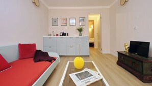 Una sala de estar moderna con un sofá, una mesa de centro con un periódico, un televisor en un mueble de madera y una cocina al fondo.