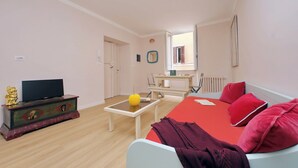Un salón con un sofá, un televisor sobre un mueble de madera, una mesa pequeña con un objeto amarillo y una ventana que da a vistas exteriores.