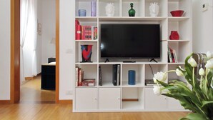A living room with a TV on a white entertainment unit, bookshelves, and a vase of flowers. - Metastasio - One Bedroom Apartment, Sleeps 4 (Rome)