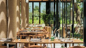 Een terras met houten tafels en stoelen, omgeven door hoge bamboeschermen en grote ramen.