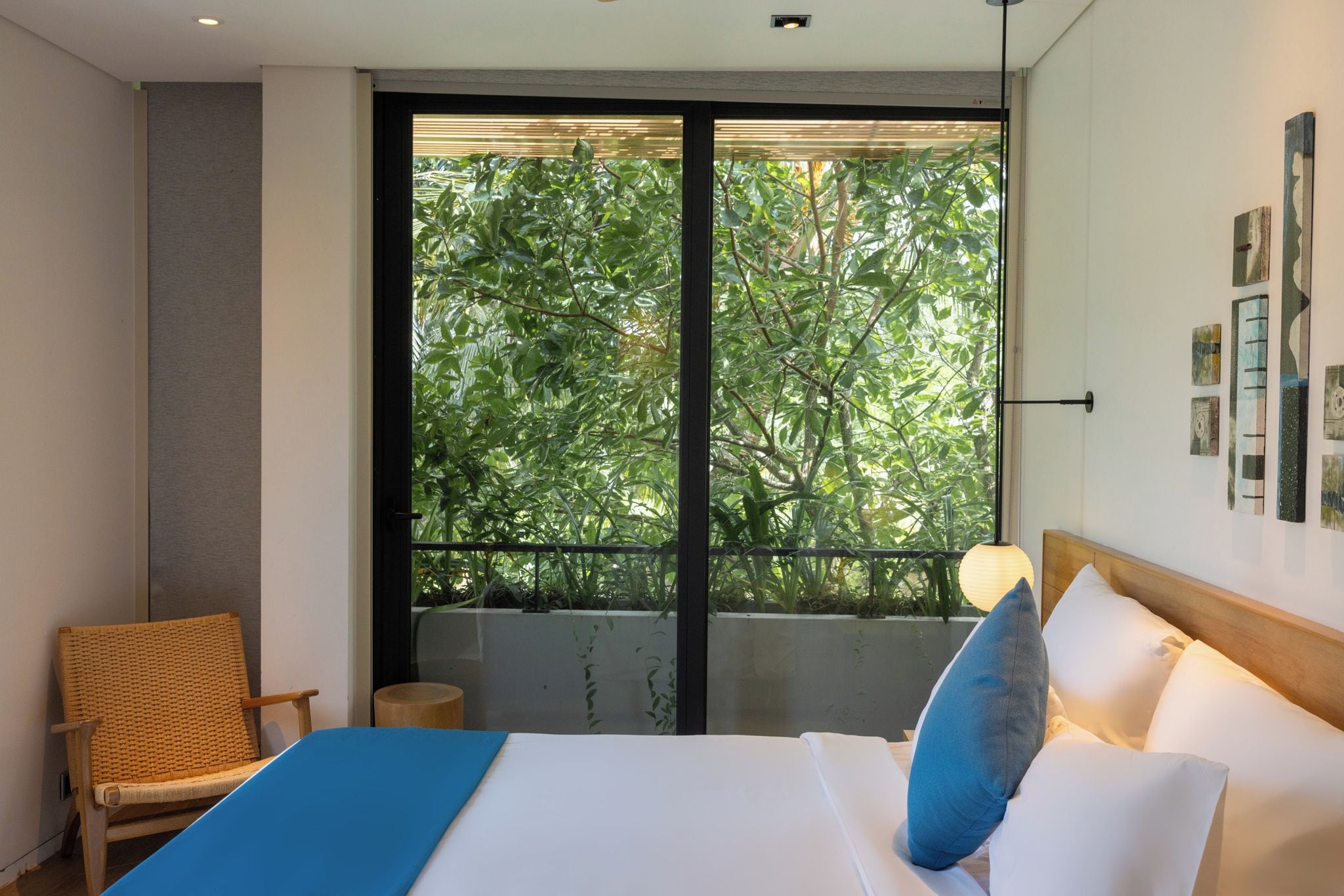 A hotel room with a bed, a chair, a view of greenery through a sliding door, and artwork on the wall.