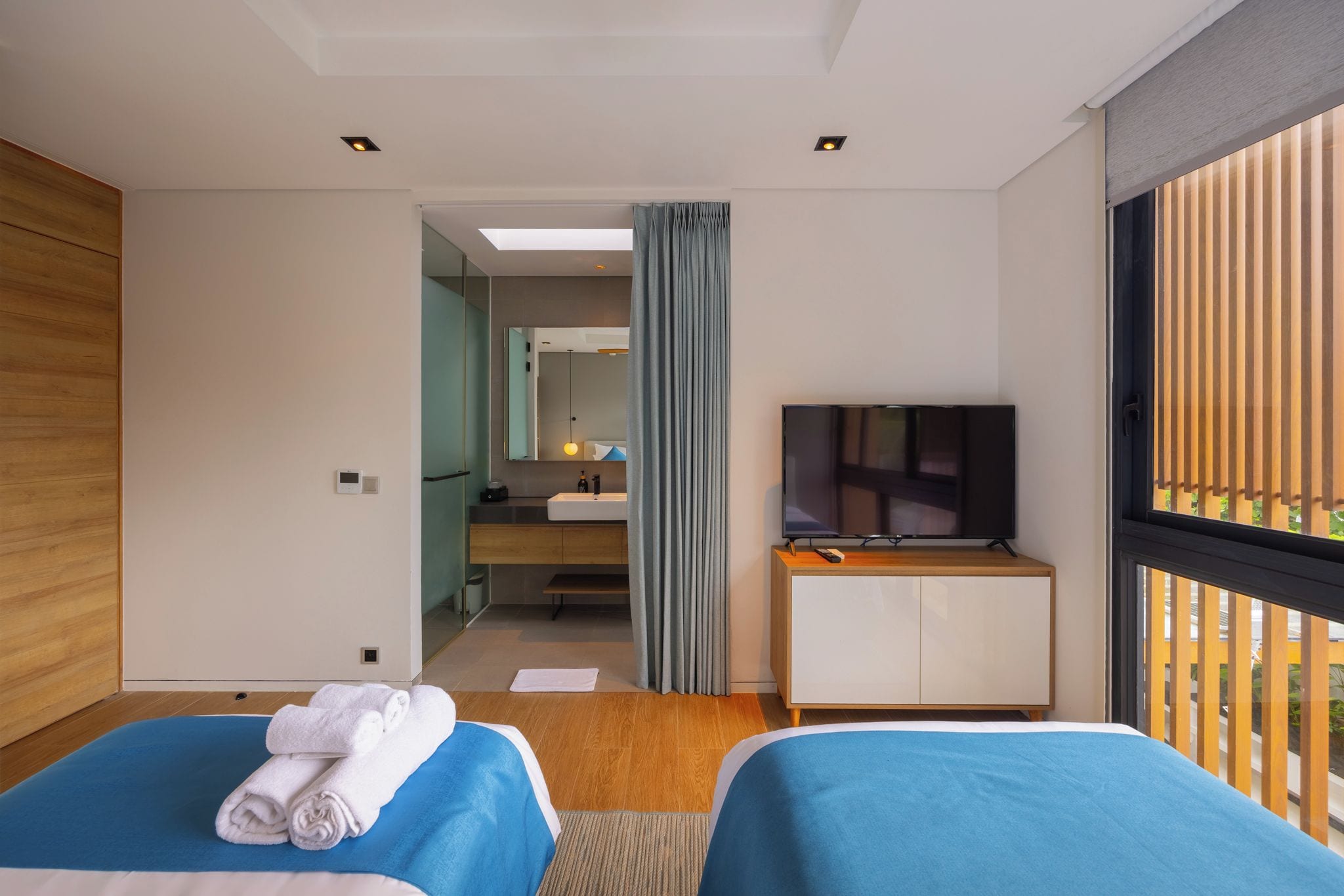 Habitación de hotel con dos camas, televisión, escritorio y baño visible a través de una puerta de cristal.