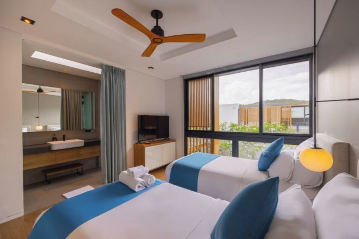 Une chambre d’hôtel avec deux lits, une télévision, un ventilateur de plafond, un balcon avec vue et une salle de bain équipée d’un lavabo et d’un miroir.