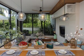 A modern dining area with a wooden table set for a meal, surrounded by large windows offering a view of lush greenery.