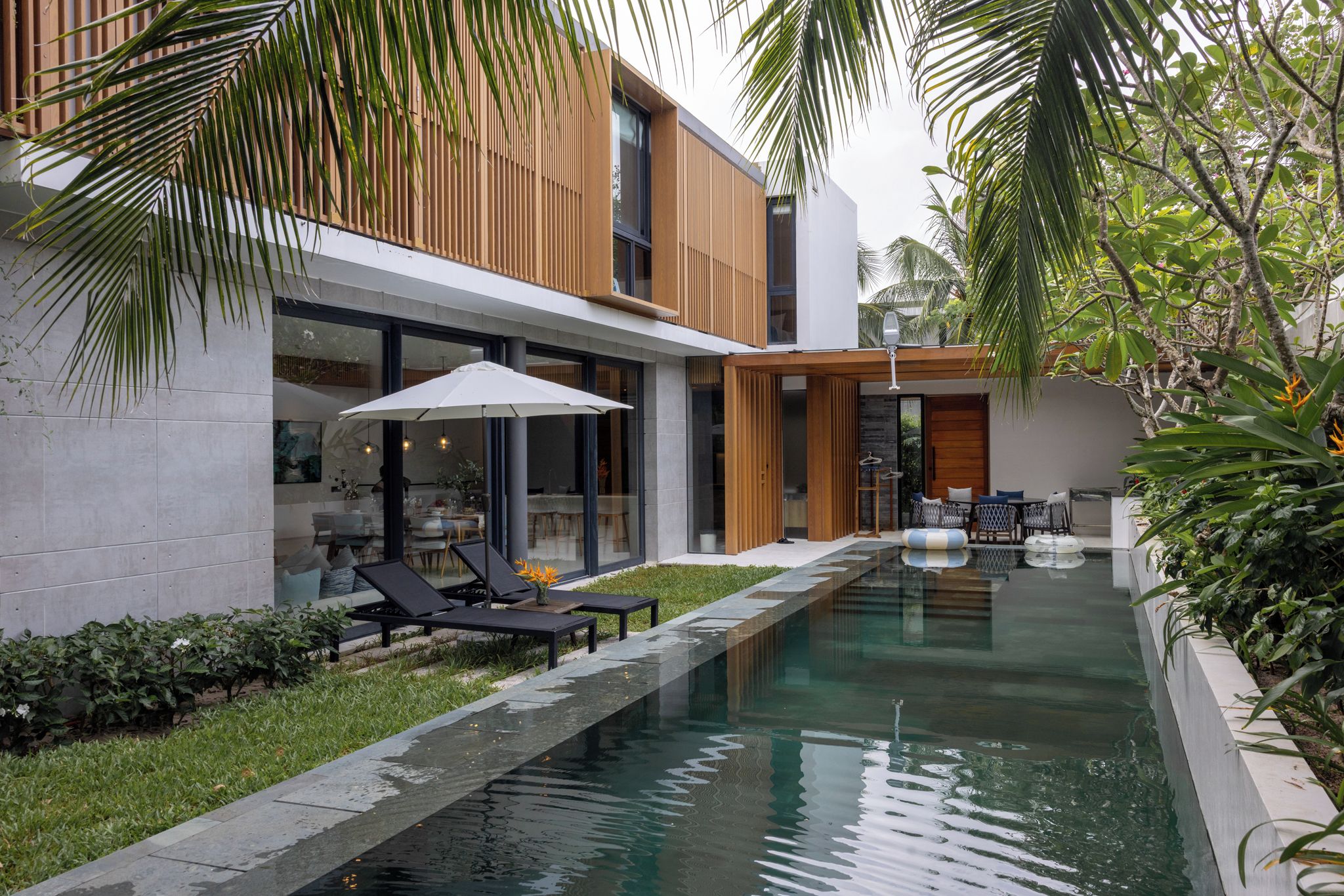 A modern outdoor pool area with lounge chairs and umbrellas, surrounded by lush greenery and a wooden deck.